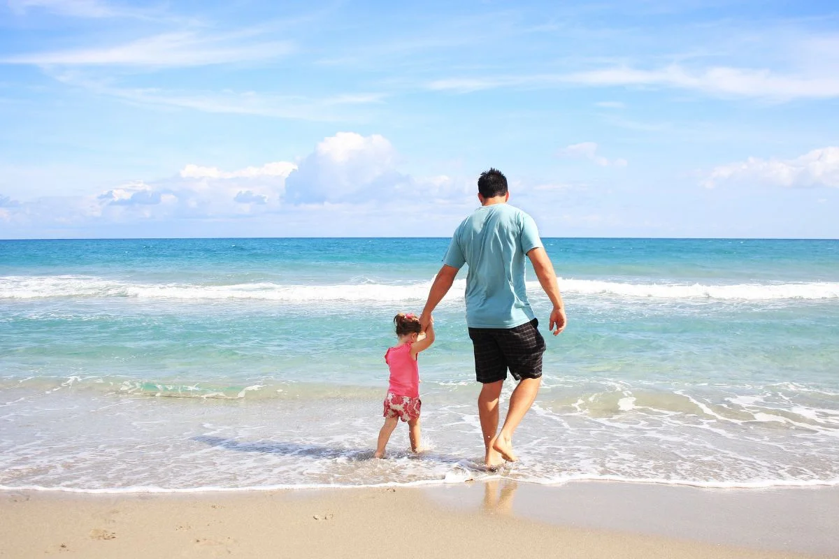 Vater geht mit seiner kleinen Tochter an der Hand ins Meer, Sinnbild für Vertrauen und Sicherheit