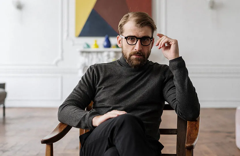 Mann mit Brille sitzt nachdenklich in einem Sessel und zweifelt an sich